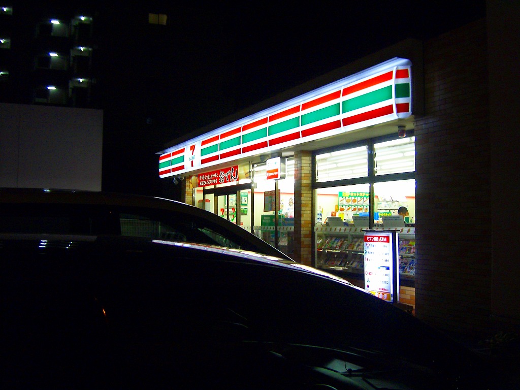暗闇に浮かぶコンビニのネオンと店内の明るい光が駐車した車を照らす